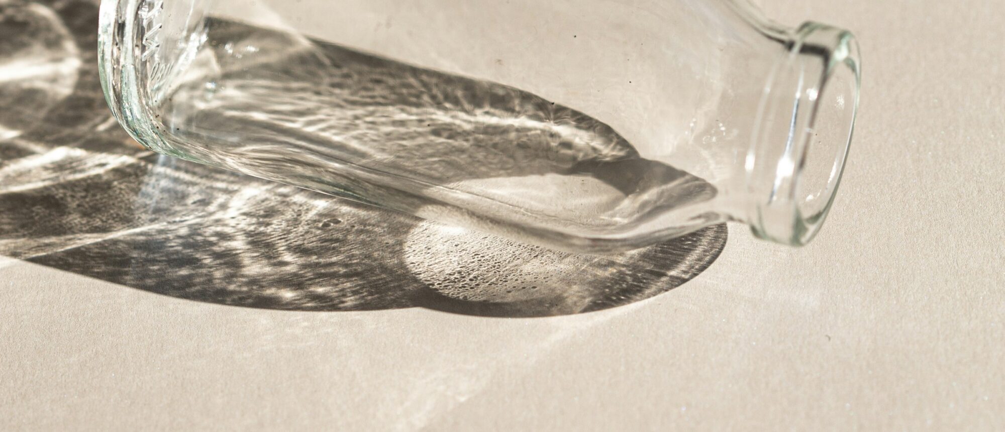 Ein gläserner Flakon liegt im Licht, Wasserreflexe auf hellem Stein